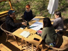 Four people sitting at a wooden table outdoors, discussing a map. There are notebooks, a bottle, and glasses on the table.