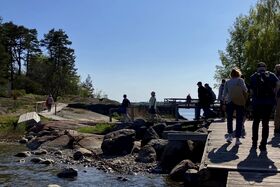 People walking on a wooden path by the rocky shore with trees in the background under a clear sky.