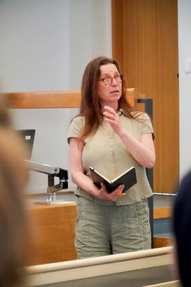 Person standing indoors, holding a notebook and a pencil, wearing a beige shirt and grey trousers.
