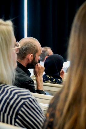 People sitting in a conference room, one wearing a striped shirt, another in a black shirt, and a person in a patterned headscarf.