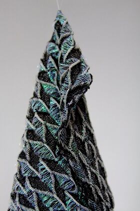 Close-up of a textured, woven fabric with black, blue, and green threads, hanging against a plain background.