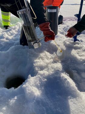 Two people collect water samples from a hole in the snow using scientific equipment. One wears orange gloves.