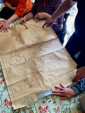 Several people examine an old map spread out on a floral tablecloth. One person points at a location on the map.