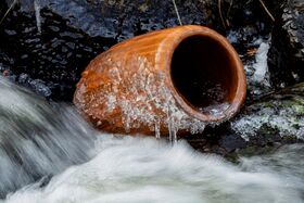 A brown clay pot partially covered in ice lies in a flowing stream surrounded by rocks.