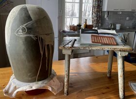 A large vase with fish drawings next to a table with an open book and pencils in a cosy kitchen.