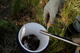 A gloved hand holds a tool over a metal pipe, with a white bucket containing soil in the background.