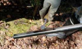 A gloved hand holds a soil sampler with soil inside, over a forest floor covered in leaves and moss.