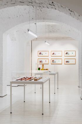 A white-walled gallery with display tables and framed artworks. The tables have glass covers showcasing various items.