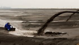 A person in a blue coat sits near a large pipe discharging dark sludge onto a barren landscape.