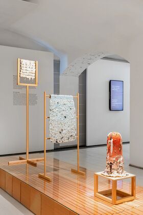 Art installation with three textured pieces on wooden stands in a modern gallery space with white walls and an arched ceiling.
