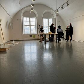 A group of people stand and sit around a table in a spacious room with large windows and grey wooden floors.