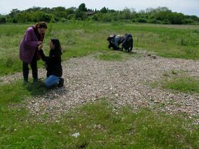 Two people are standing and kneeling on a grassy field with scattered pebbles. Another person is crouching in the background.