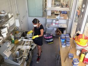 A person wearing yellow gloves works in a cluttered kitchen with various containers and equipment.