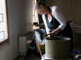 A person wearing a black apron is shaping a large clay pot in a room with a window, heater, and various items on a chair.