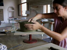 A person shaping clay on a rotating stand in a studio with tables, tools, and a window in the background.