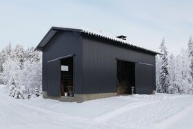 A dark grey barn with open doors stands in a snowy landscape surrounded by snow-covered trees.