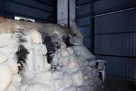 A stone kiln with blackened areas inside a metal building. A white plastic chair is on the right.