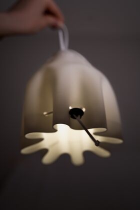A hand holding a white, modern pendant lamp with a unique, wavy design. The lamp is lit, casting a soft glow.