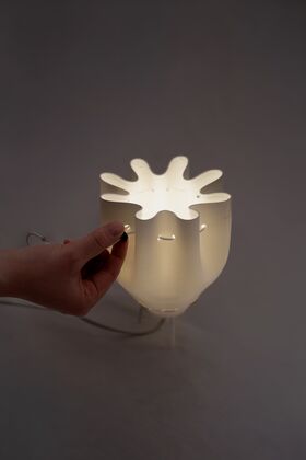 A hand adjusts a modern white lamp with a unique, wavy design. The lamp emits a soft glow in a dimly lit room.