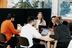 Five people sitting at a table with laptops, engaged in discussion. Plants and large windows are in the background.