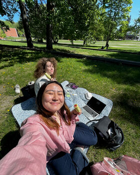 Two people having a picnic on a blanket with a laptop, bags, and food in a park with trees and grass.