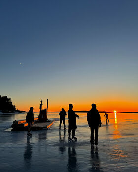 People ice skating on a frozen lake at sunset. A small boat is docked nearby. The sky is clear with a gradient from blue to orange.