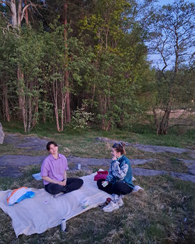 Two people sitting on a blanket in a grassy area surrounded by trees. They have snacks and drinks with them.