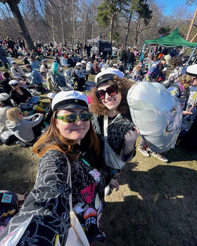 Two people in white student caps holding a balloon at an outdoor event. Many people are sitting on the grass under trees.