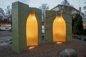 Two large stone sculptures with bottle-shaped cutouts illuminated from inside, situated in a gravel area near a building.