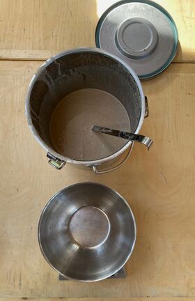 Two metal containers on a wooden surface, one with a ladle and brown liquid, the other empty. A lid is partially visible.