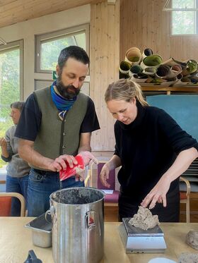 Two people working with clay in a room with large windows and wooden walls. Rolled-up mats are visible in the background.
