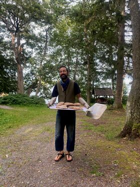 A person holding a tray with freshly baked bread outdoors, surrounded by trees and greenery.