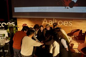 A group of people stand in a circle with their hands joined in the centre. Aaltoes sign is visible in the background.