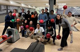 A group of people at a bowling alley holding bowling balls, posing near the lanes and ball return machine.