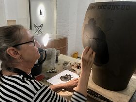 A person is drawing a detailed insect on a large vase in a studio. The workspace has a lamp, papers, and art supplies.