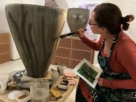 A person painting a large vase with a brush, holding a reference image. Art supplies are scattered around.