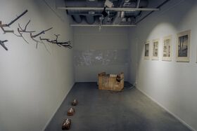 A gallery room with branches on the left wall, framed pictures on the right wall, and a wooden crate in the centre.