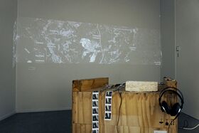 A map is projected on a wall above a wooden table with headphones and a strip of photos.