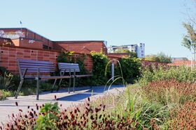 Kaksi penkkiä päällystetyllä polulla tiiliseinän vieressä, jossa on vihreää kasvillisuutta. Taustalla näkyy rakennuksia.