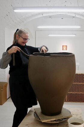 A person working on a large clay pot in a studio with white walls and bright overhead lights.
