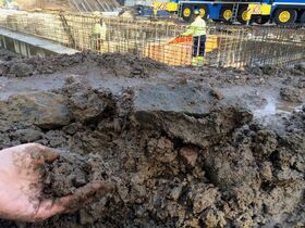 A muddy construction site with a worker in a yellow jacket and helmet. A hand holds a clump of wet soil in the foreground.