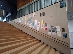 Wide wooden stairs in a modern building, with a beige wall covered in colourful posters along the railing.