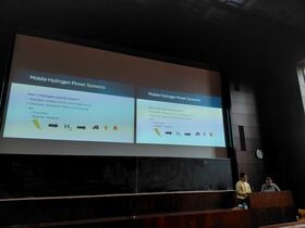 Kaksi esittäjää hämärässä luentosalissa näyttää diaslidean liikkuvista vetyöljyjärjestelmistä.