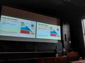 Luentosalissa kolme ihmistä esittelee kaksinkertaisesti heijastettuja energiasäilytysdioja taulun yläpuolella.