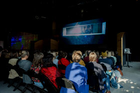 People are sitting and watching a movie trailer on a large screen.