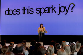 A person speaks at a podium in front of a large screen displaying 'does this spark joy?'.