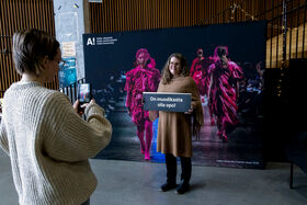 Henkilö pitää kylttiä, toinen ottaa valokuvaa. Taustalla on muotinäytös.