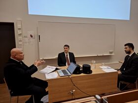 Three men in suits discuss at a lectern with laptop, papers and two black academic hats in a classroom