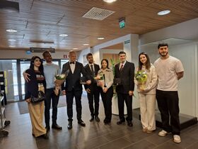 Seven people indoors in smart clothing, some holding flower bouquets, posing together in a lobby-like space