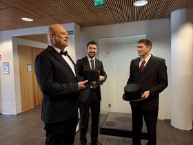 Three men in dark suits indoors, holding black cylindrical academic hats in front of a glass display.